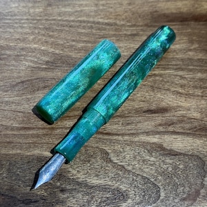 Op de afbeelding: Een vulpen met een glinsterend groen en blauw gemarmerd ontwerp. De dop is eraf en toont de penpunt. De pen ligt op een houten oppervlak.