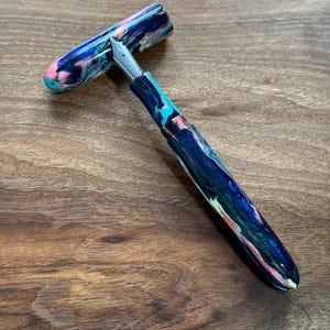 May include: A colorful fountain pen with a marbled design in shades of blue, pink, and green. The pen features a silver nib and a matching cap, lying on a wooden surface. The pen is a writing instrument.