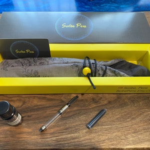 May include: A pen set in a yellow box with the text "Scribe Pens". The set includes a clear pen, ink bottle, and a floral patterned pen wrap. The box also contains a business card with the "Scribe Pens" logo.