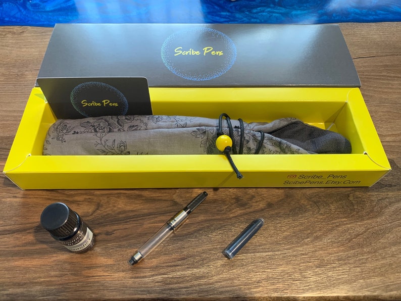 May include: A pen set in a yellow box with the text "Scribe Pens". The set includes a clear pen, ink bottle, and a floral patterned pen wrap. The box also contains a business card with the "Scribe Pens" logo.