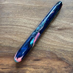 May include: A handcrafted fountain pen with a marbled design. The pen features a dark blue base color with swirls of pink, teal, and white. The pen is shown on a wooden surface.