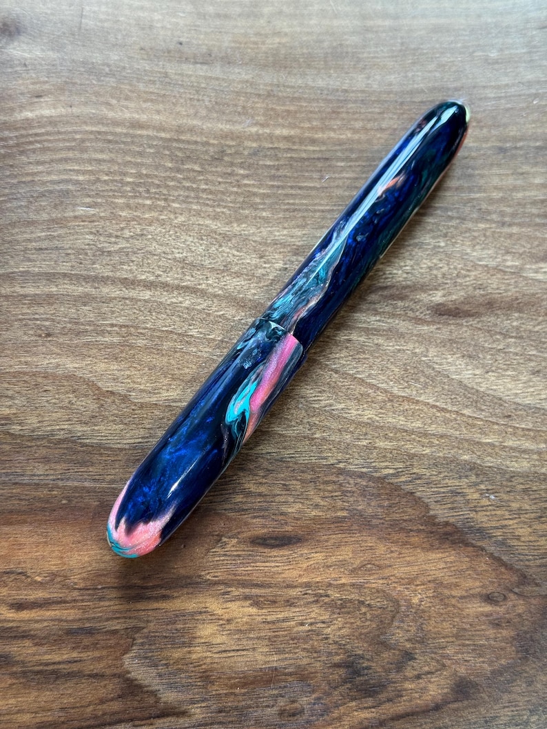 May include: A handcrafted fountain pen with a marbled design. The pen features a dark blue base color with swirls of pink, teal, and white. The pen is shown on a wooden surface.