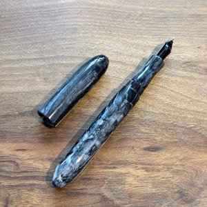 May include: A fountain pen with a marbled black and white design, with the cap removed and placed to the side. The pen has a black nib and a sleek, modern design. The pen is lying on a wooden surface.