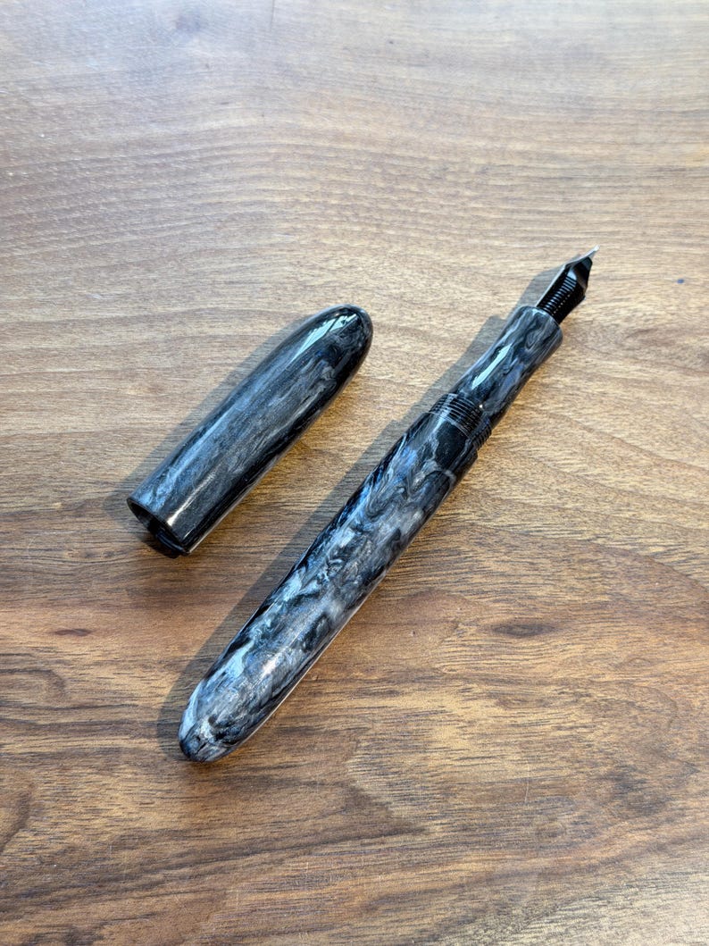 May include: A fountain pen with a marbled black and white design, with the cap removed and placed to the side. The pen has a black nib and a sleek, modern design. The pen is lying on a wooden surface.