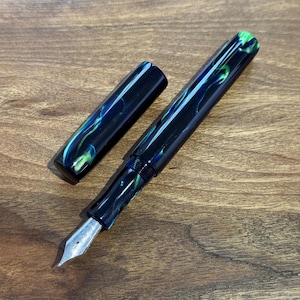 May include: A fountain pen with a silver nib and a black barrel with blue and green swirls. The cap is off to the side, revealing the pen's design. The pen is lying on a wooden surface.