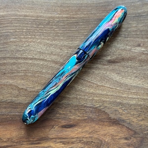 May include: A colorful fountain pen with a marbled design. The pen features a mix of blue, teal, pink, and green hues. The pen's body has a glossy finish and a black band near the center. The pen is lying on a wooden surface.