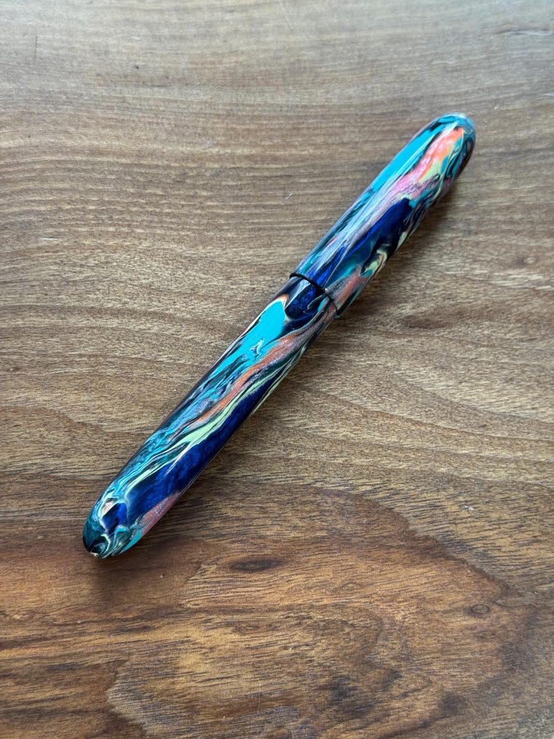 May include: A colorful fountain pen with a marbled design. The pen features a mix of blue, teal, pink, and green hues. The pen's body has a glossy finish and a black band near the center. The pen is lying on a wooden surface.