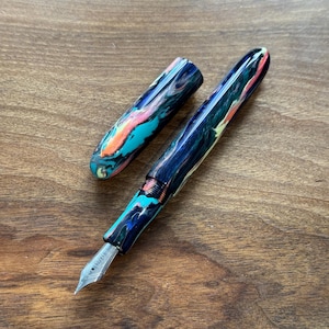 May include: A colorful fountain pen with a marbled design in shades of blue, teal, pink, and orange. The pen is open, with the cap placed to the side, revealing the silver nib. The pen rests on a wooden surface.
