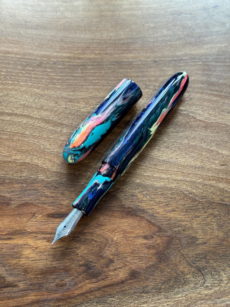 May include: A colorful fountain pen with a marbled design in shades of blue, teal, pink, and orange. The pen is open, with the cap placed to the side, revealing the silver nib. The pen rests on a wooden surface.