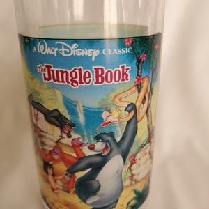 以下が含まれることがあります： ディズニーの「ジャングル・ブック」のカラフルなイラストが描かれた透明なグラス。 バルー熊やカー蛇などのキャラクターがジャングルの背景に描かれています。「A Walt Disney Classic」と「The Jungle Book」の文字が目立ちます。
