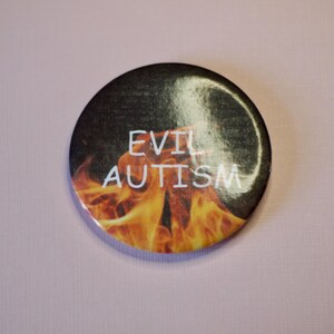 以下が含まれることがあります： 炎を背景に「EVIL AUTISM」の文字が白で書かれた丸いボタン。オレンジと黄色の炎のデザインが施された、黒とグレーの背景です。