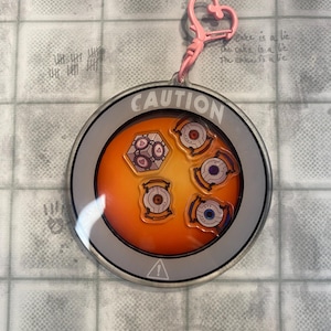 Può includere: Un portachiavi rotondo con un fermaglio rosa a forma di cuore. Il portachiavi ha un bordo grigio con la scritta "CAUTION" e un simbolo di avvertimento. All'interno, uno sfondo arancione con disegni di robot.