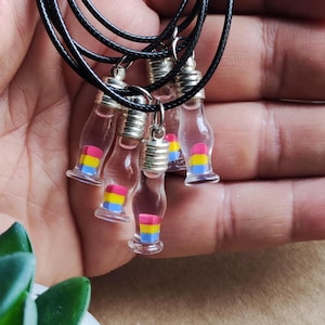 Pode incluir: Cinco colares com pequenos frascos de vidro contendo uma bandeira do orgulho pansexual. Os frascos estão presos a cordões pretos.
