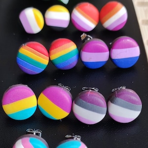 Könnte beinhalten: Eine Sammlung bunter Ohrstecker mit verschiedenen LGBTQ+-Pride-Flaggen-Designs. Die Ohrringe zeigen Regenbogen-, lesbische, nicht-binäre, bisexuelle, pansexuelle, asexuelle und Transgender-Pride-Flaggen. Die Ohrringe sind rund und haben silberne Stifte.