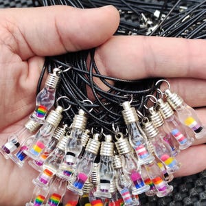 Può includere: Una collezione di collane con piccole bottiglie di vetro che contengono diverse bandiere arcobaleno. Le bottiglie sono attaccate a cordini neri.