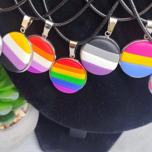 Op de afbeelding: Een set van acht kettingen met verschillende pride-vlag hangers. De hangers zijn rond en hebben verschillende kleuren en ontwerpen, waaronder regenboog-, transgender-, biseksuele- en panseksuele vlaggen.