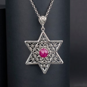 Könnte beinhalten: Eine silberne Kettenkette mit einem Anhänger in Form eines Davidsterns. Der Anhänger ist filigran gearbeitet und hat einen rosafarbenen Edelstein in der Mitte.