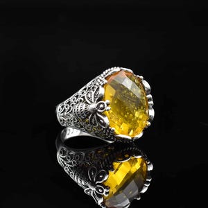 May include: A silver ring with an oval, yellow gemstone. The ring has an ornate, filigree design around the band and setting. The gemstone is faceted and reflects light, creating a sparkling effect. The ring is displayed on a reflective surface.