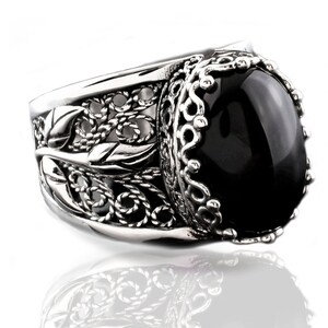 May include: A silver ring with an ornate design featuring a black oval gemstone. The ring has a filigree pattern with leaves and swirls.