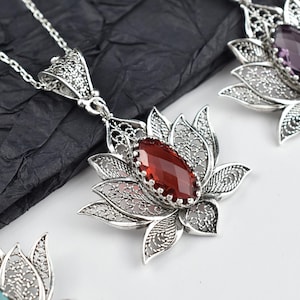 May include: Silver filigree lotus flower pendant necklace with a faceted, oval, red gemstone center. The pendant is suspended from a delicate silver chain. Other necklaces with different colored gemstones are also visible.