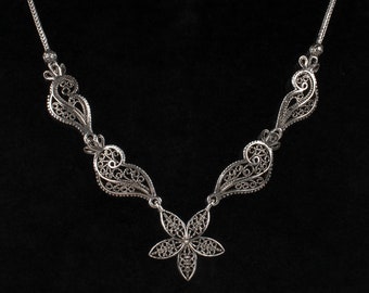 May include: A silver filigree necklace with a delicate flower pendant. The necklace is made of intricate, interwoven silver wire, creating a delicate and elegant design.