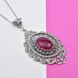 May include: A silver filigree pendant necklace with a large, oval, faceted, ruby-colored gemstone. The pendant has an ornate, openwork design and hangs from a delicate chain. The background is white and pink.