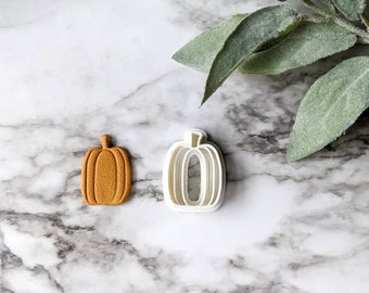 Può includere: Un tagliapasta a forma di zucca in plastica bianca e un biscotto di zucchero marrone a forma di zucca. Gli articoli sono su una superficie di marmo bianco.