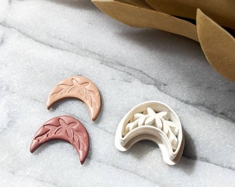 Könnte beinhalten: Zwei sichelförmige Ausstecher, einer aus weißem Kunststoff und der andere aus hellbraunem Ton. Beide haben ein florales Design.