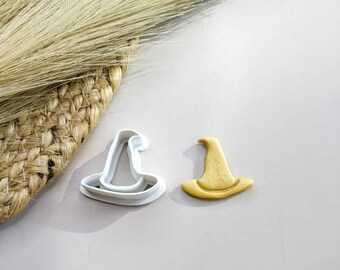 Può includere: Un tagliabiscotti bianco a forma di cappello da strega accanto a un biscotto giallo cotto a forma di cappello da strega. Il tagliabiscotti è su una superficie grigio chiaro. L'immagine ha uno sfondo neutro.
