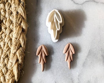 Peut inclure: Deux boucles d'oreilles en argile brunes en forme de feuille et un emporte-pièce en plastique blanc en forme de feuille sur une surface en marbre blanc.