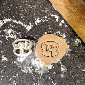 Peut inclure: Un emporte-pièce en plastique blanc en forme de maison avec le mot "Luke's" à l'intérieur, sur une surface grise avec un rouleau à pâtisserie et une découpe de pâte à biscuits brune.
