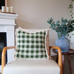 以下が含まれることがあります： 白の肘掛け椅子の上に、緑と白のギンガムチェック柄のクロシェ編みクッションが置かれています。クッションは白いクロシェ編みの縁取りがされています。肘掛け椅子は暖炉と緑色の花瓶のあるリビングルームに置かれています。