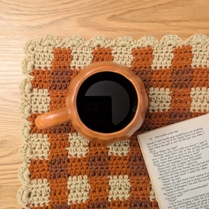 Può includere: Vista dall'alto di una tazza di ceramica marrone piena di liquido nero, appoggiata su un sottobicchiere all'uncinetto fatto a mano. Il sottobicchiere presenta un motivo a scacchi nei toni del marrone e del beige.