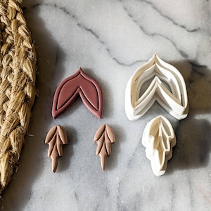 Pode incluir: Cinco cortadores de biscoitos em diferentes formas e tamanhos. Dois são marrons, dois são brancos e um é de um tom marrom mais escuro. Os cortadores estão sobre uma superfície de mármore branco.