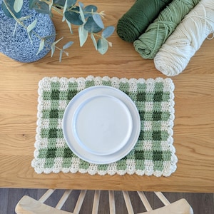 May include: A green and white gingham crochet placemat with a scalloped edge and a white plate on top.