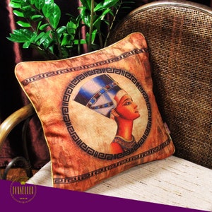 May include: Decorative throw pillow featuring an illustration of an Egyptian queen. The pillow has a brown background with a circular design around the queen's portrait. The queen is depicted with a blue headdress and red skin. The pillow is on a chair.