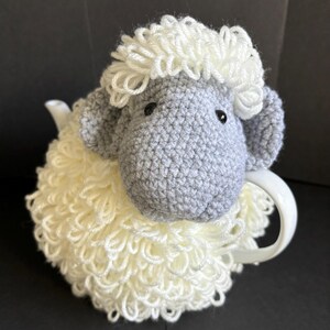 May include: A crocheted gray and white sheep tea cozy. The sheep has a fluffy white fleece and a gray face with black eyes.