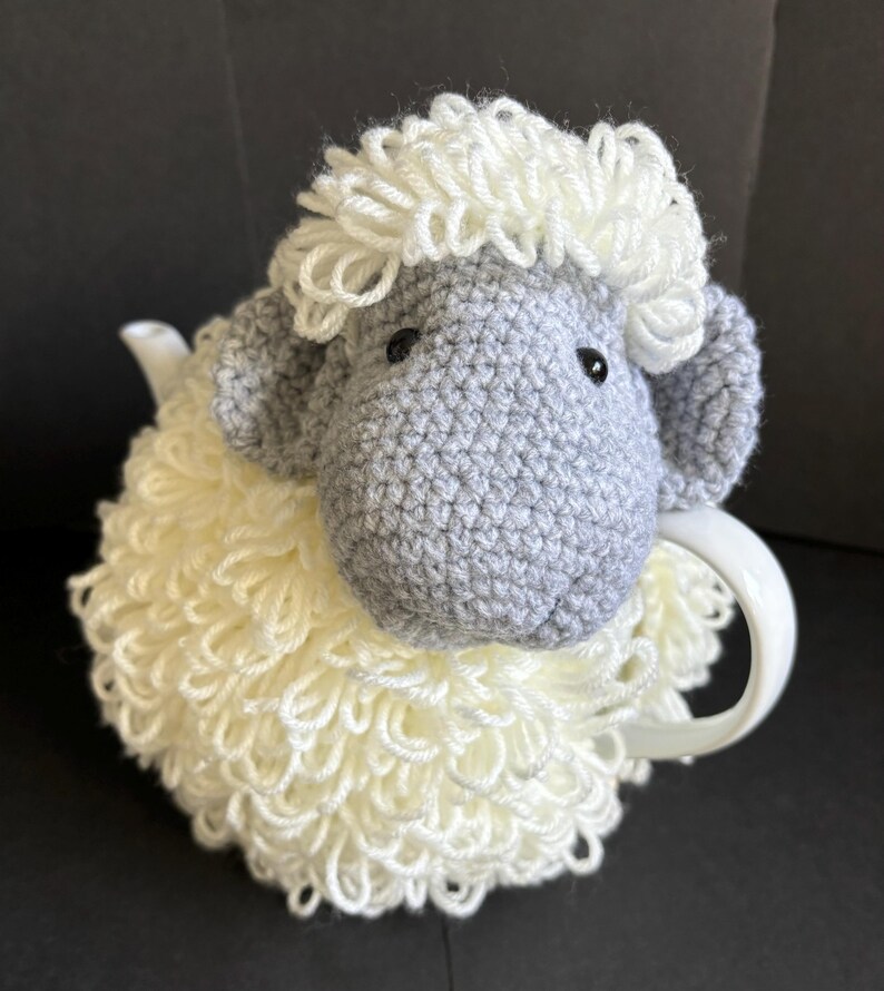 May include: A crocheted gray and white sheep tea cozy. The sheep has a fluffy white fleece and a gray face with black eyes.