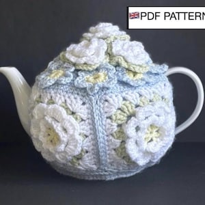 Puede incluir: Una tetera de cerámica blanca cubierta con un cubretejera de ganchillo. El cubretejera es azul claro, blanco y amarillo pálido, con grandes aplicaciones de flores blancas. El texto "PDF PATTERN" es visible.