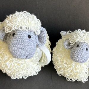 May include: Two crocheted sheep tea cozy sets. The larger set is gray and white and the smaller set is white. Both sets have a fluffy white wool texture.