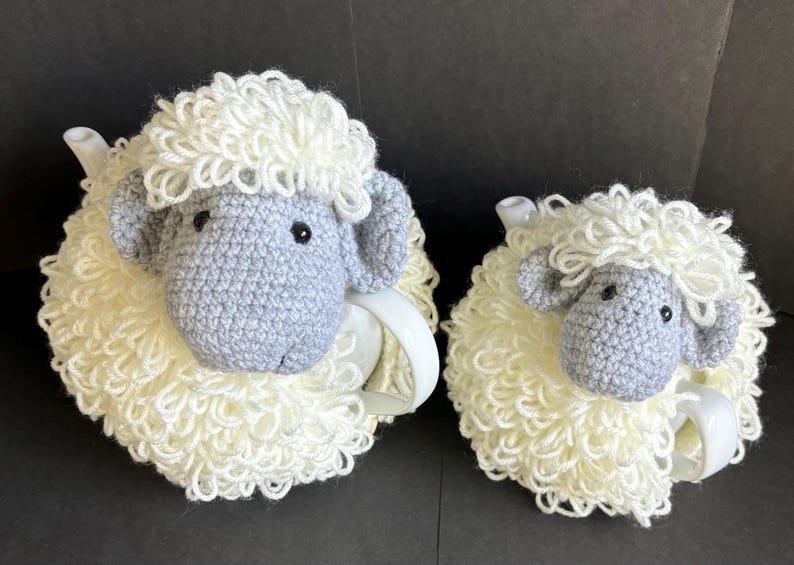 May include: Two crocheted sheep tea cozy sets. The larger set is gray and white and the smaller set is white. Both sets have a fluffy white wool texture.