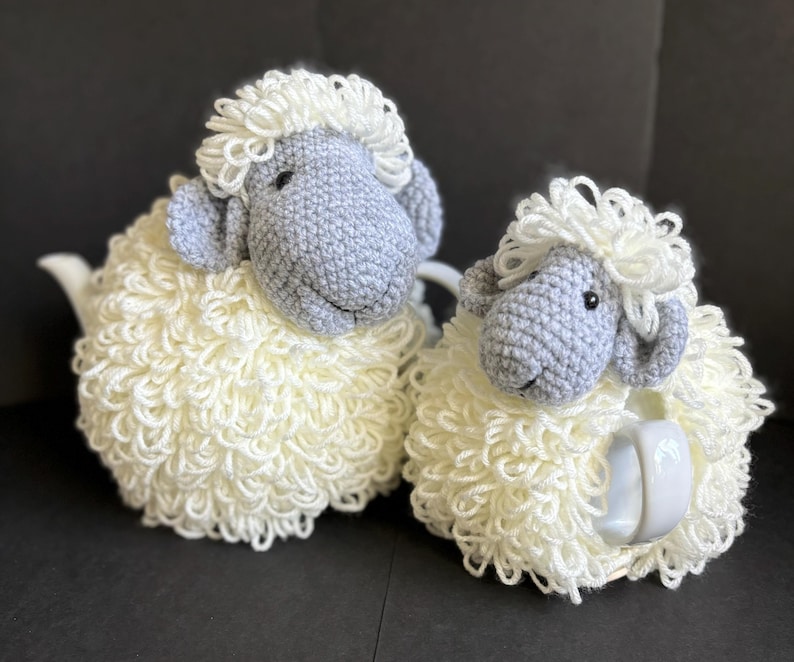 May include: Two crocheted sheep tea cozy sets. The larger sheep is white with a gray face and the smaller sheep is white with a gray face and a white teapot.