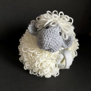 May include: A crocheted white sheep mug cozy with a gray sheep head and black eyes. The cozy is wrapped around a white mug.