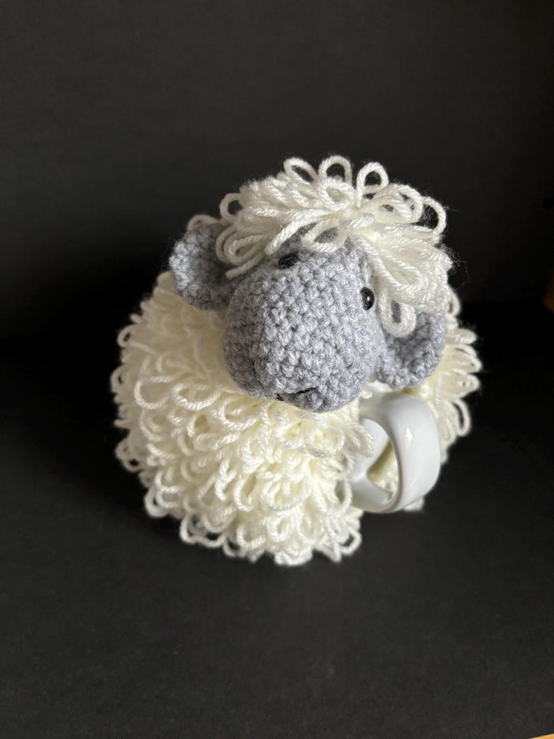 May include: A crocheted white sheep mug cozy with a gray sheep head and black eyes. The cozy is wrapped around a white mug.