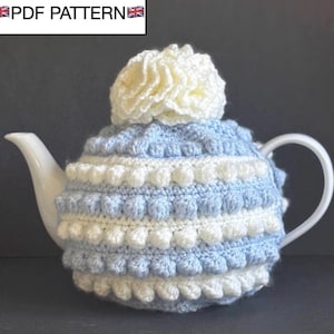 Puede incluir: Una tetera de cerámica blanca cubierta con un cubrete de ganchillo azul y blanco. El cubrete tiene un diseño en capas con una gran flor de color crema en la parte superior. El texto "PDF PATTERN" está en la esquina superior izquierda.