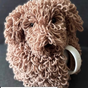 Könnte beinhalten: Eine handgefertigte, hellbraune, hundeförmige Tasse aus Garn. Die Tasse hat schwarze Augen und eine weiße Tasse ist teilweise sichtbar. Der Text "PDF PATTERN" befindet sich am unteren Rand des Bildes.