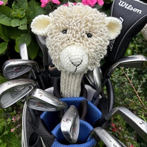 May include: A golf club bag with a selection of clubs, topped with a cream-coloured sheep headcover. The bag is black and blue, set against a backdrop of green foliage and pink flowers. A Wilson club cover is also visible.