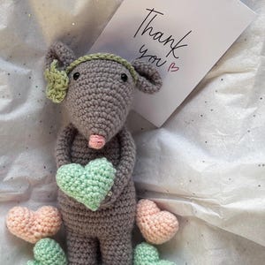 May include: A crocheted gray mouse doll holding a green heart, surrounded by pink and green crocheted hearts. The mouse has a green headband and a pink nose. A card with "Thank you" written on it is in the background.