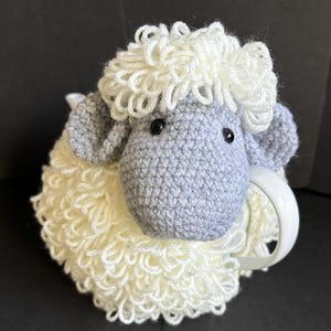 May include: A crocheted gray and white sheep mug cozy. The sheep has a fluffy white wool body and a gray face with black eyes.