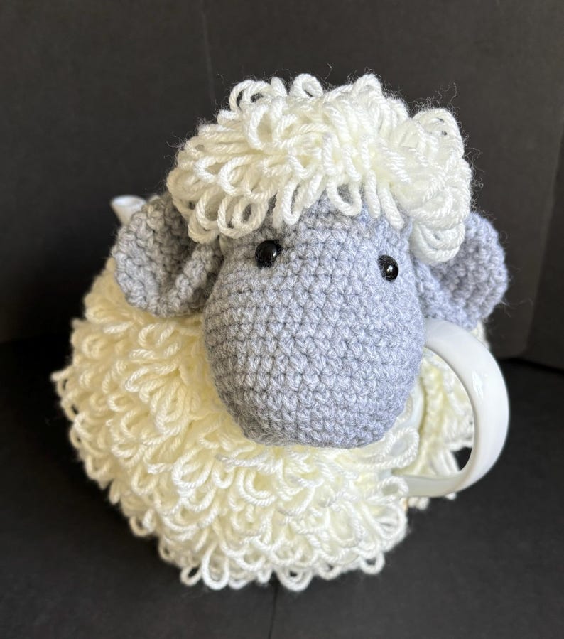 May include: A crocheted gray and white sheep mug cozy. The sheep has a fluffy white wool body and a gray face with black eyes.